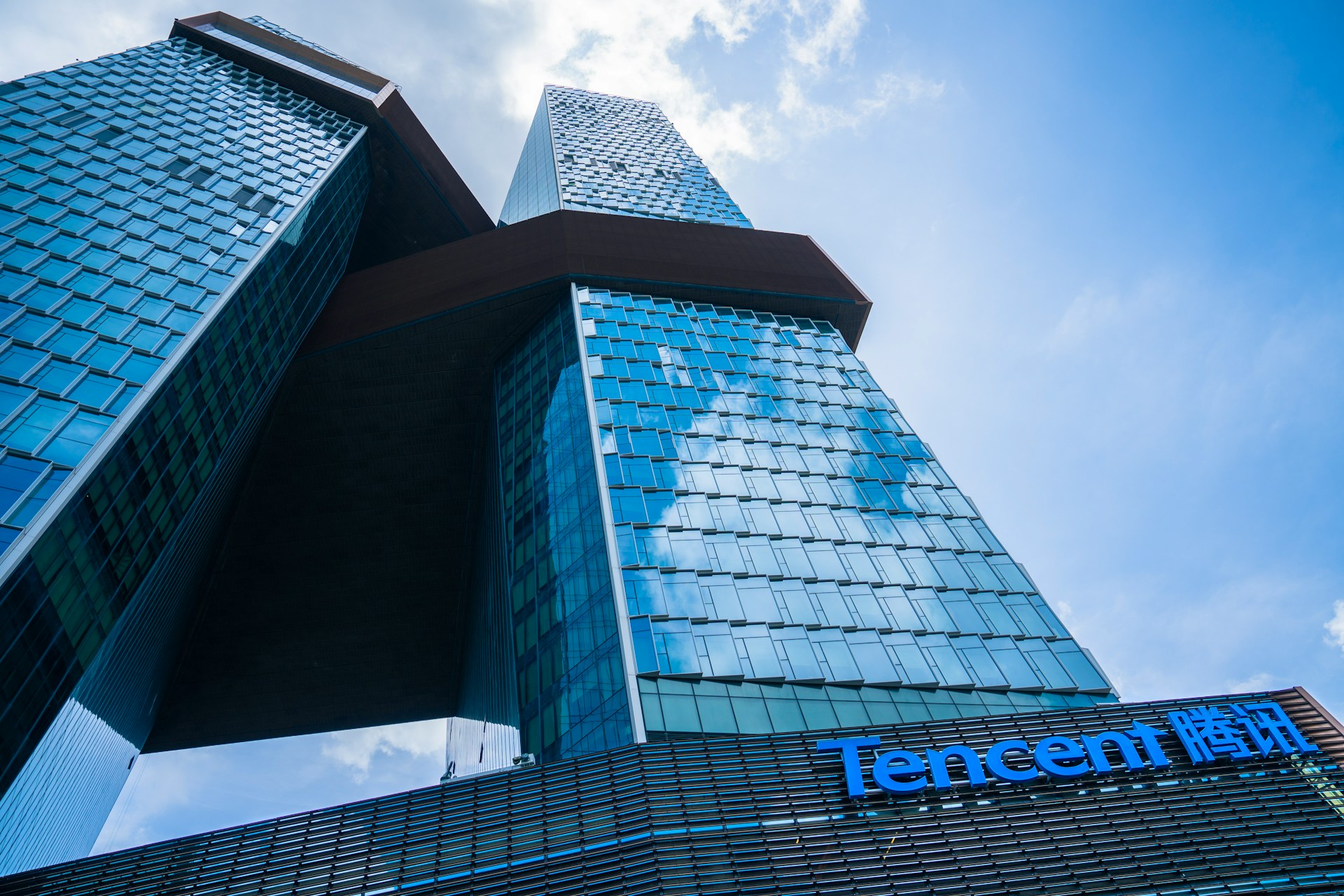 Mavi gökyüzünde dağınık bulutların önünde, mavi cam cepheye sahip ve üzerinde "Tencent" logosu belirgin şekilde yer alan yüksek, modern bir bina.